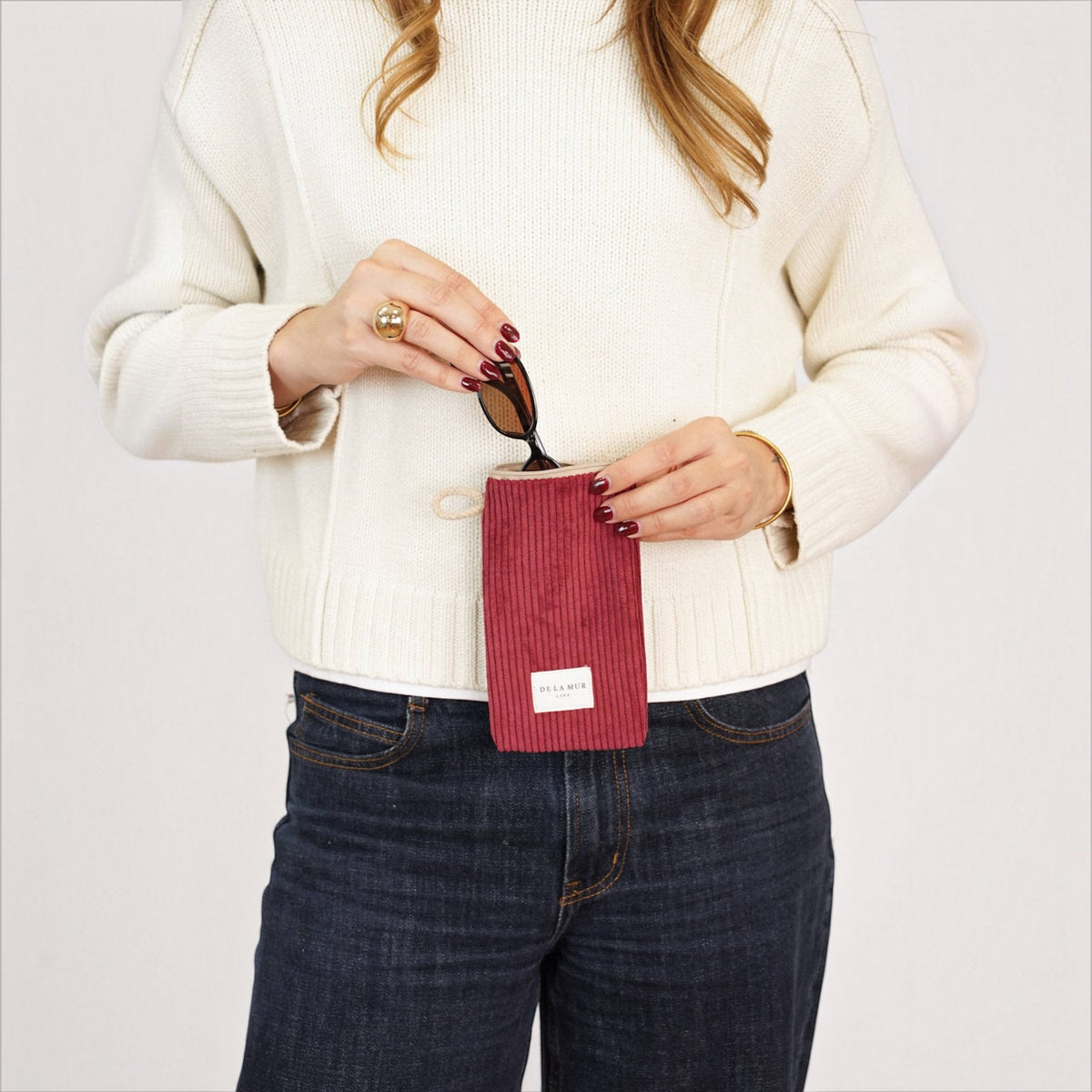 Connemara Country burgundy corduroy glasses case with zipper and beige cord handle held by a woman in a white sweater.