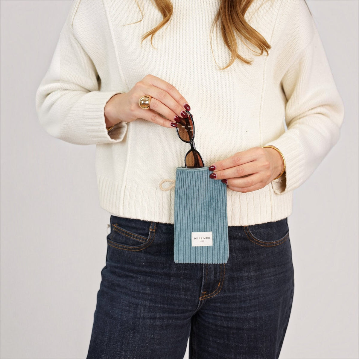 Connemara Country blue corduroy glasses case with zipper and beige cord handle held by a woman in a white sweater.
